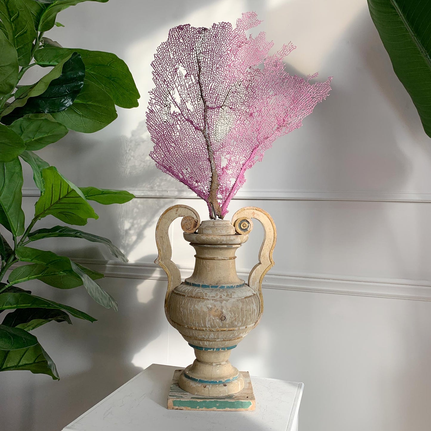 A decorative pink coral reef arrangement displayed in a distressed antique beige urn-shaped vase with two handles, set on a white surface against a white paneled wall with green plants at the sides.