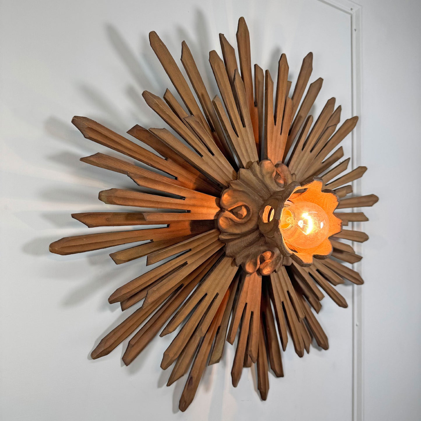 A decorative wall-mounted sunburst light fixture made of carved wood rays, with a warm glowing bulb inside a flower-shaped glass shade, shown against a plain white wall.