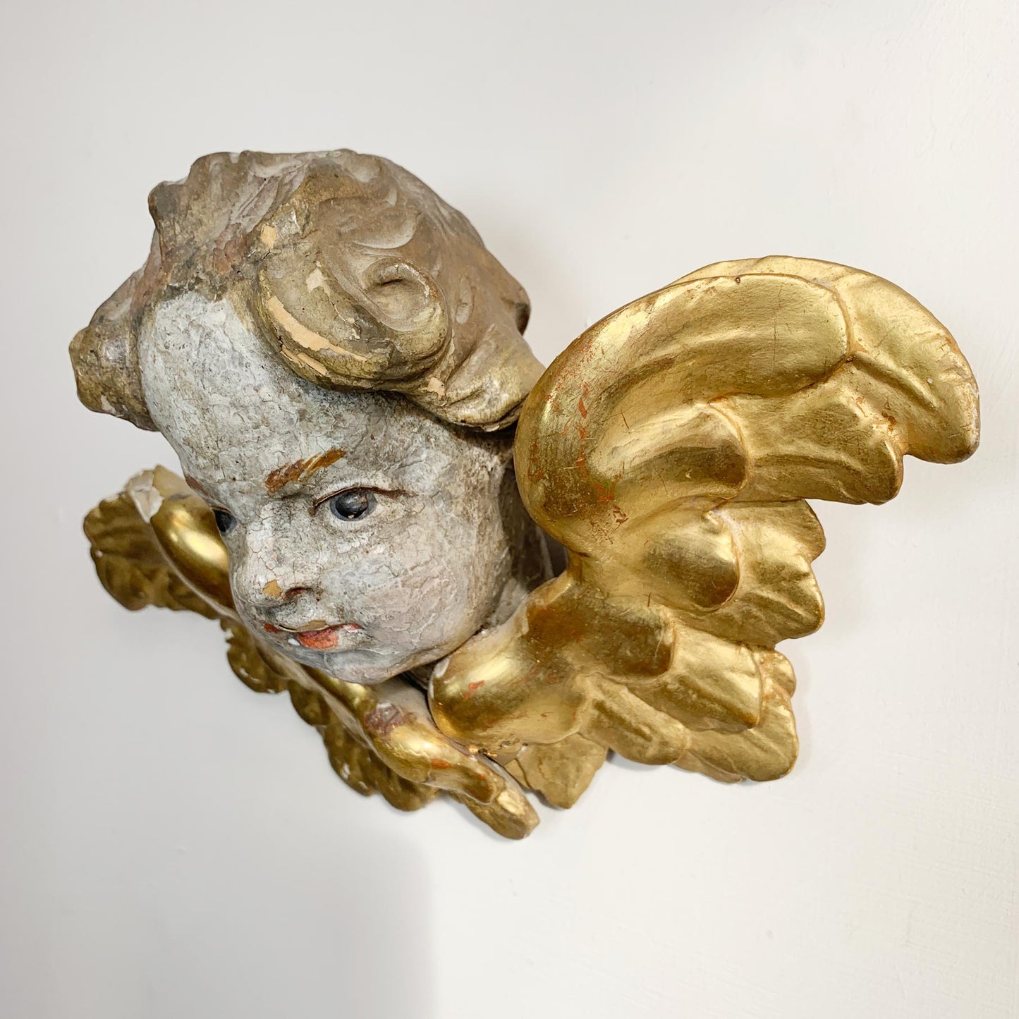 An antique gilded cherub wall sconce featuring a painted face with distressed gray and white tones, detailed with ornate gold wings and scrollwork.
