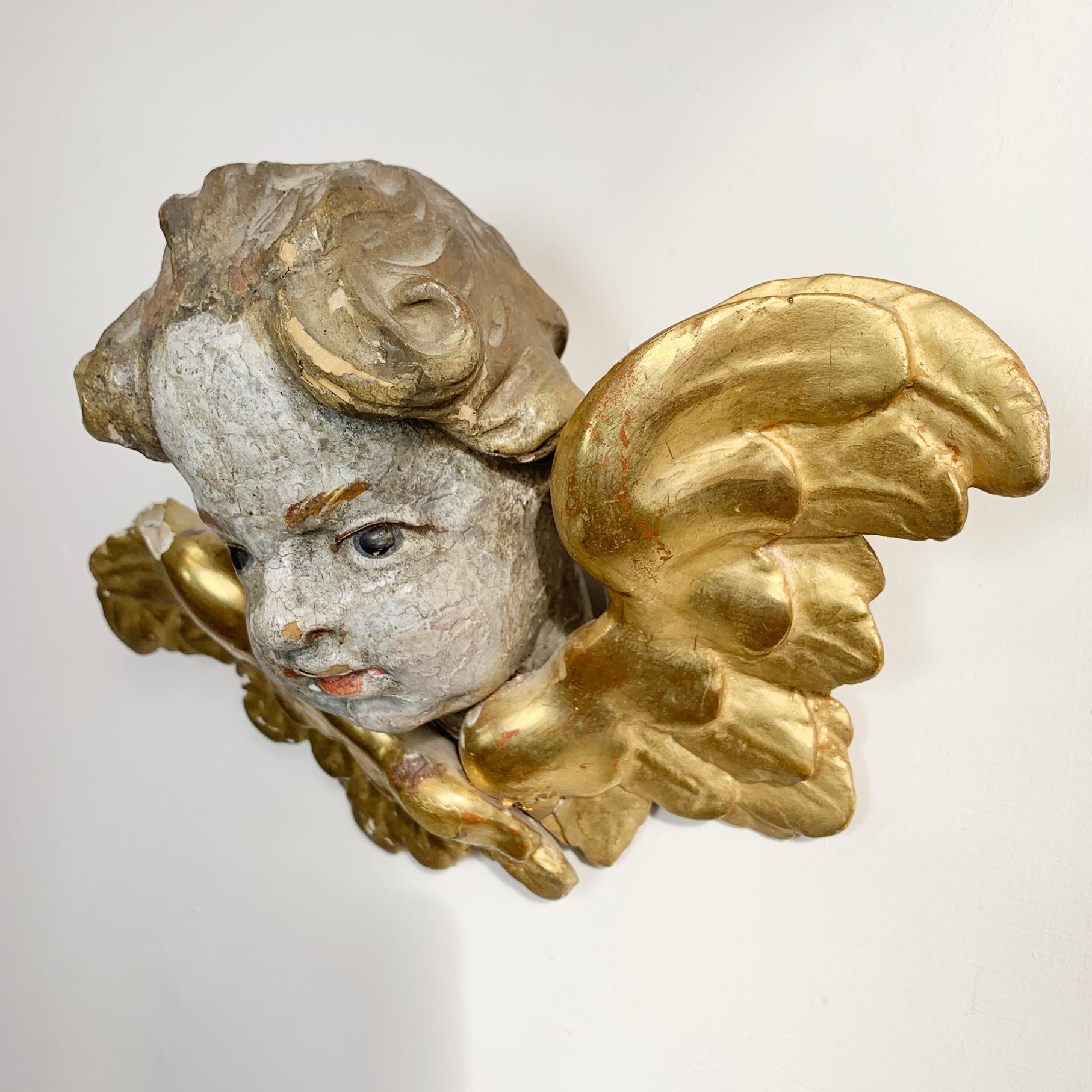 An antique gilded cherub wall sconce featuring a painted face with distressed gray and white tones, detailed with ornate gold wings and scrollwork.