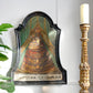 An antique religious wall plaque depicting the Coronation of Jesus Christ, mounted on a white wall next to a gold candlestick and a green plant.