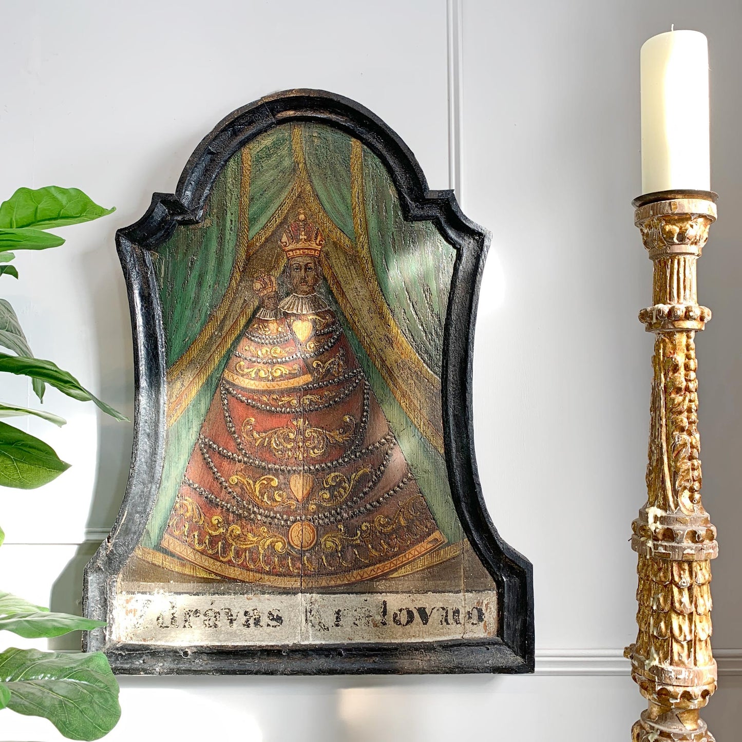 An antique religious wall plaque depicting the Coronation of Jesus Christ, mounted on a white wall next to a gold candlestick and a green plant.