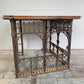 An antique ecclesiastical table featuring a rustic wooden oak top and an ornate dark cast iron frame with scrollwork and decorative brackets, set against a white wall and weathered floor.