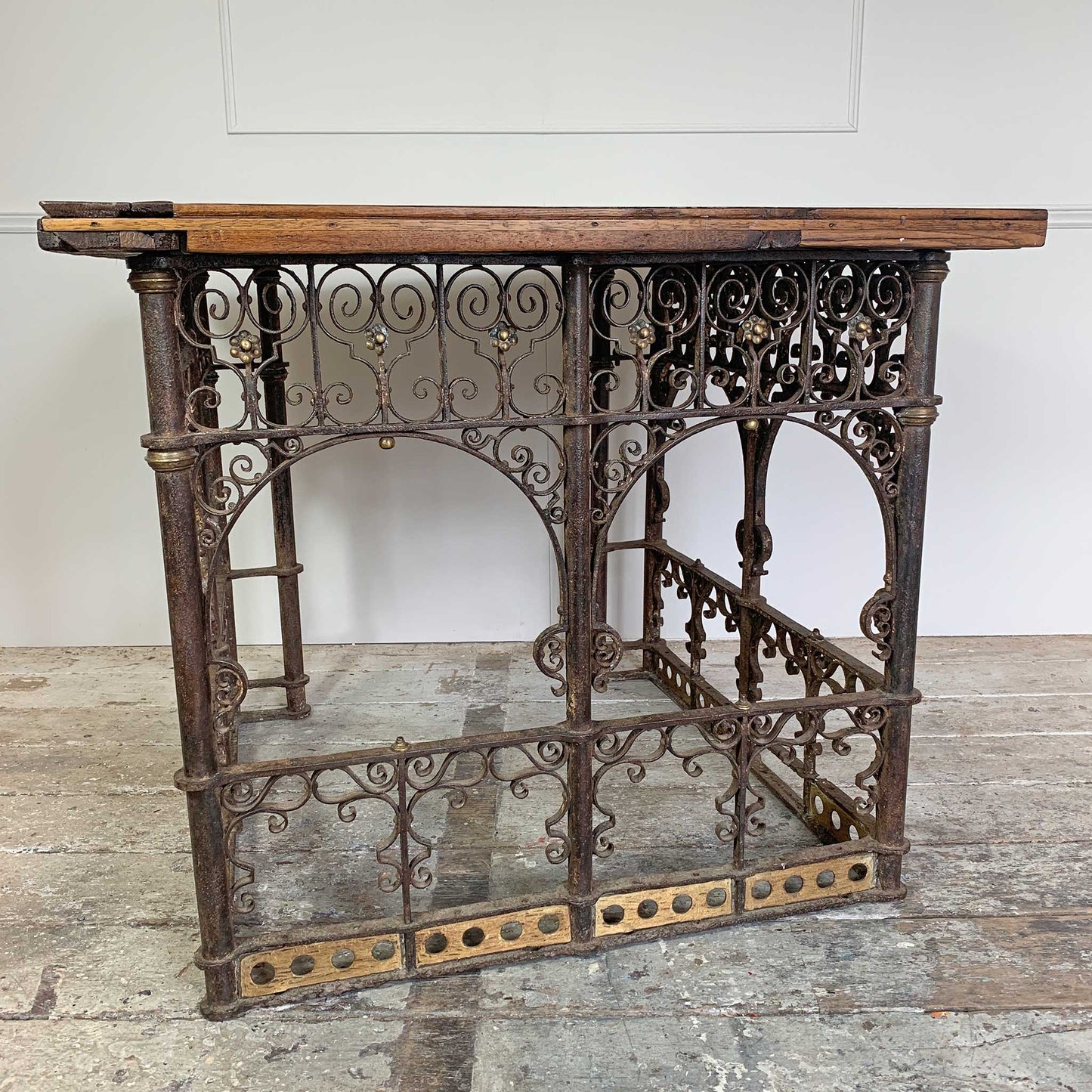 An antique ecclesiastical table featuring a rustic wooden oak top and an ornate dark cast iron frame with scrollwork and decorative brackets, set against a white wall and weathered floor.