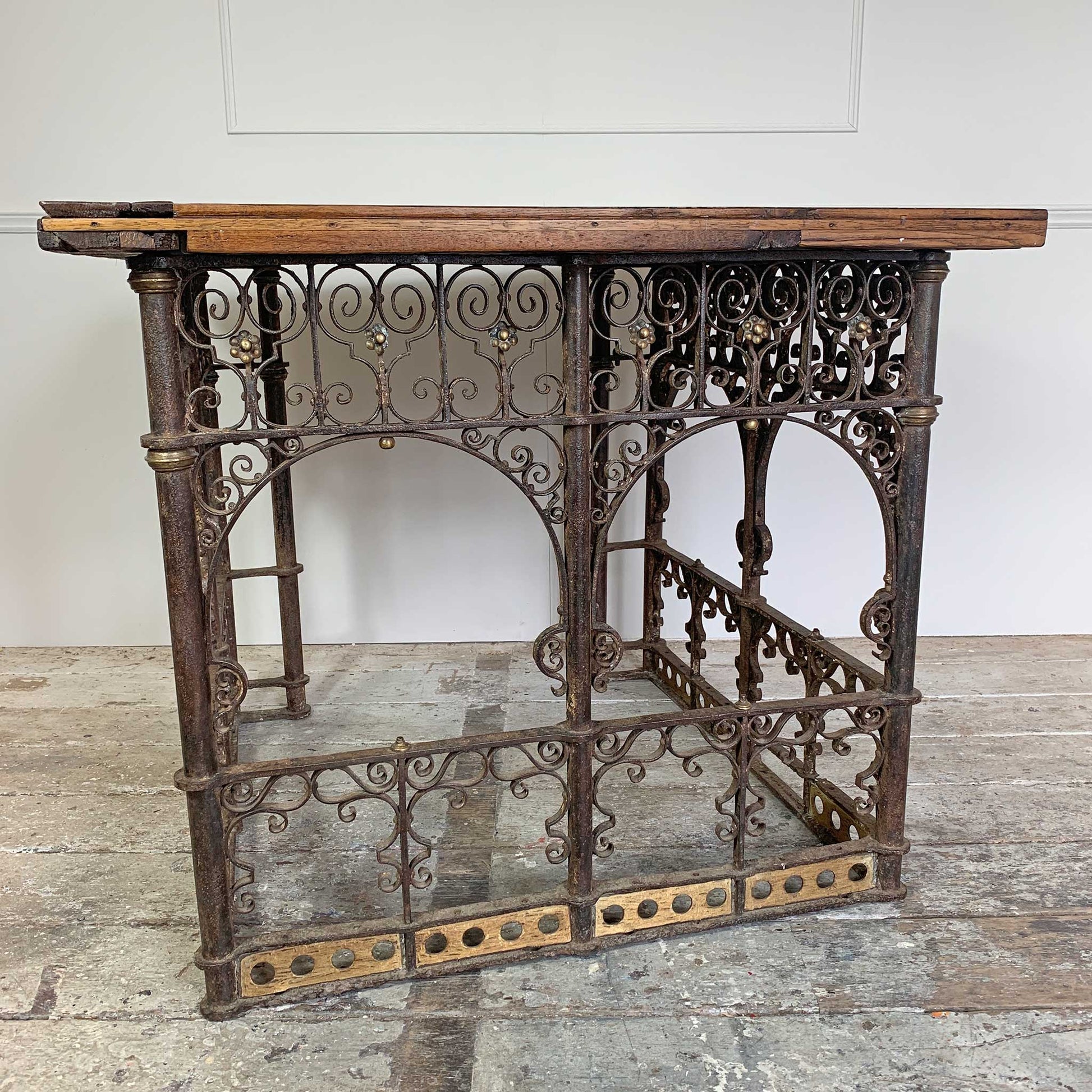 An antique ecclesiastical table featuring a rustic wooden oak top and an ornate dark cast iron frame with scrollwork and decorative brackets, set against a white wall and weathered floor.