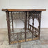 An antique ecclesiastical table featuring a rustic wooden oak top and an ornate dark cast iron frame with scrollwork and decorative brackets, set against a white wall and weathered floor.