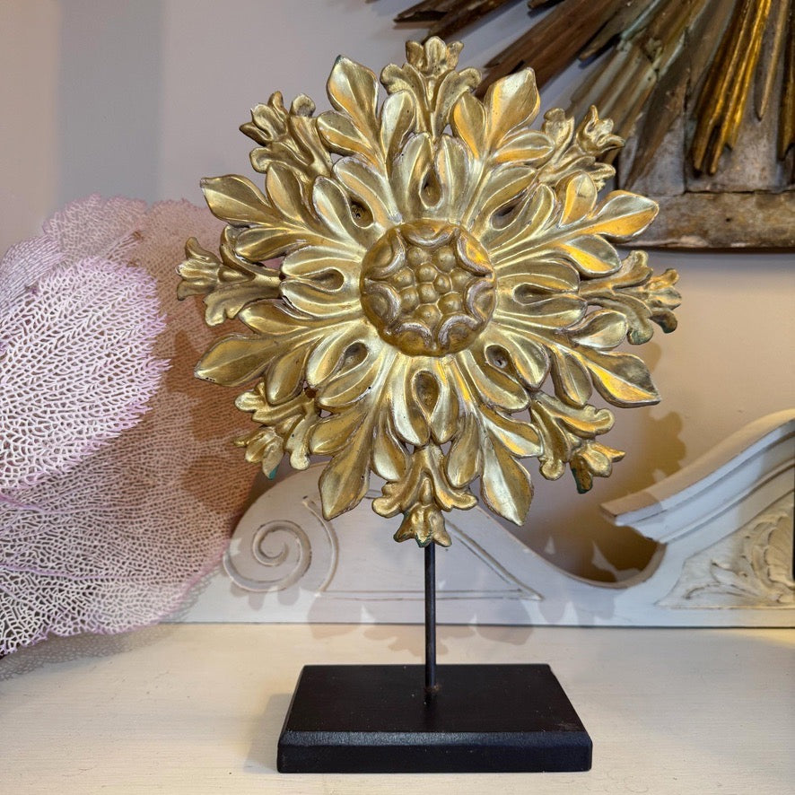 Antique gilt wooden hand carved flower sunburst wall ornament with a black metal stand, displayed on a tabletop against a softly blurred interior background.