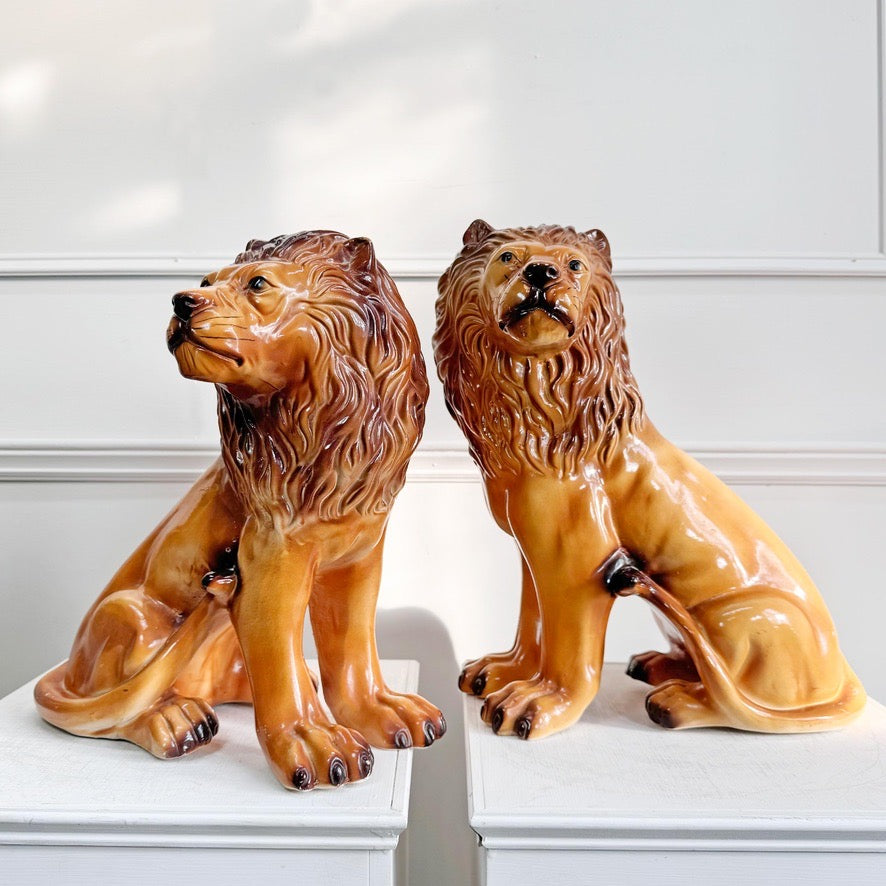 Two glossy ceramic Staffordshire lion figurines in warm tan and brown tones, posed side by side on white pedestals against a white paneled wall.