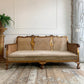 An antique French-style three-seater sofa with a carved gold-leaf wooden frame, woven cane backrest, and beige velvet seat cushion, set in a rustic room with a wooden floor and a potted fern on the left.
