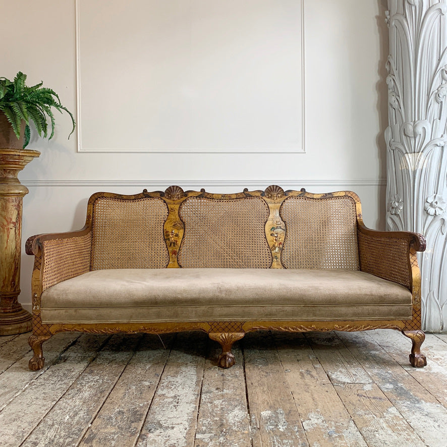 An antique French-style three-seater sofa with a carved gold-leaf wooden frame, woven cane backrest, and beige velvet seat cushion, set in a rustic room with a wooden floor and a potted fern on the left.