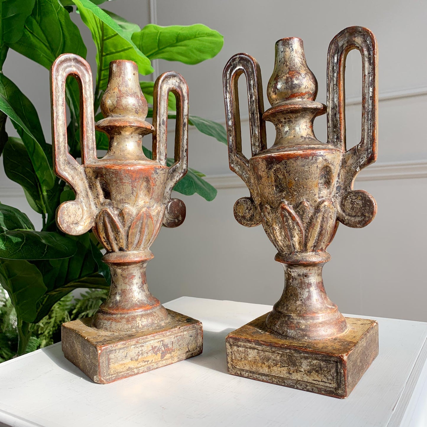 A pair of 18th century decorative urn bookends with a distressed bronze and gold finish, displayed on a white surface with green leaves in the background.