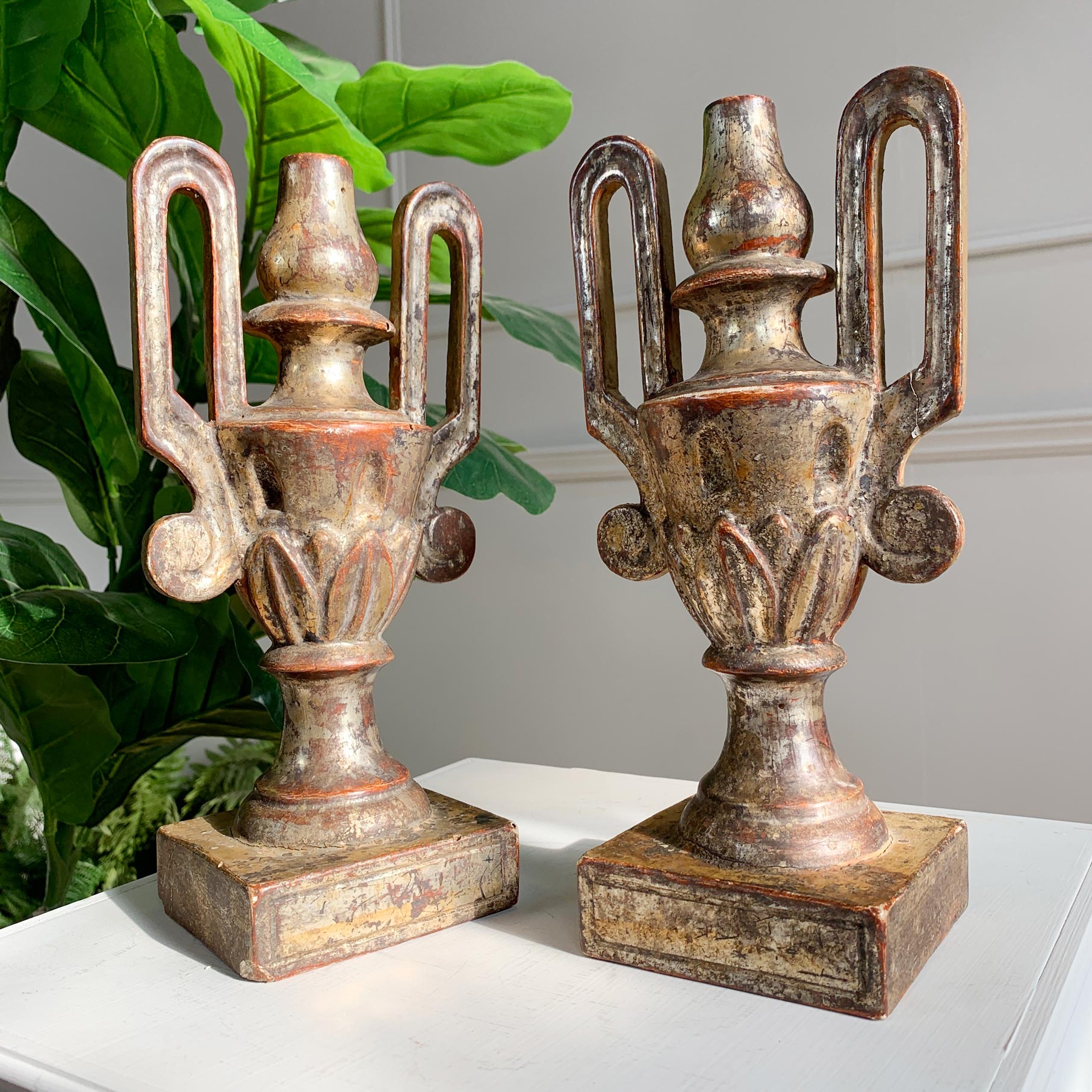 A pair of 18th century decorative urn bookends with a distressed bronze and gold finish, displayed on a white surface with green leaves in the background.