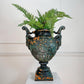 An antique cast iron Durenne urn planter with a distressed green and orange patina, filled with green ferns, displayed on a white pedestal against a white paneled wall.