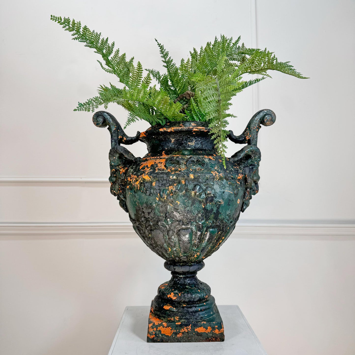 An antique cast iron Durenne urn planter with a distressed green and orange patina, filled with green ferns, displayed on a white pedestal against a white paneled wall.