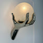 A sculptural maison arlus wall sconce featuring a glossy metallic hand holding a glowing, crackled glass orb against a plain light gray wall.