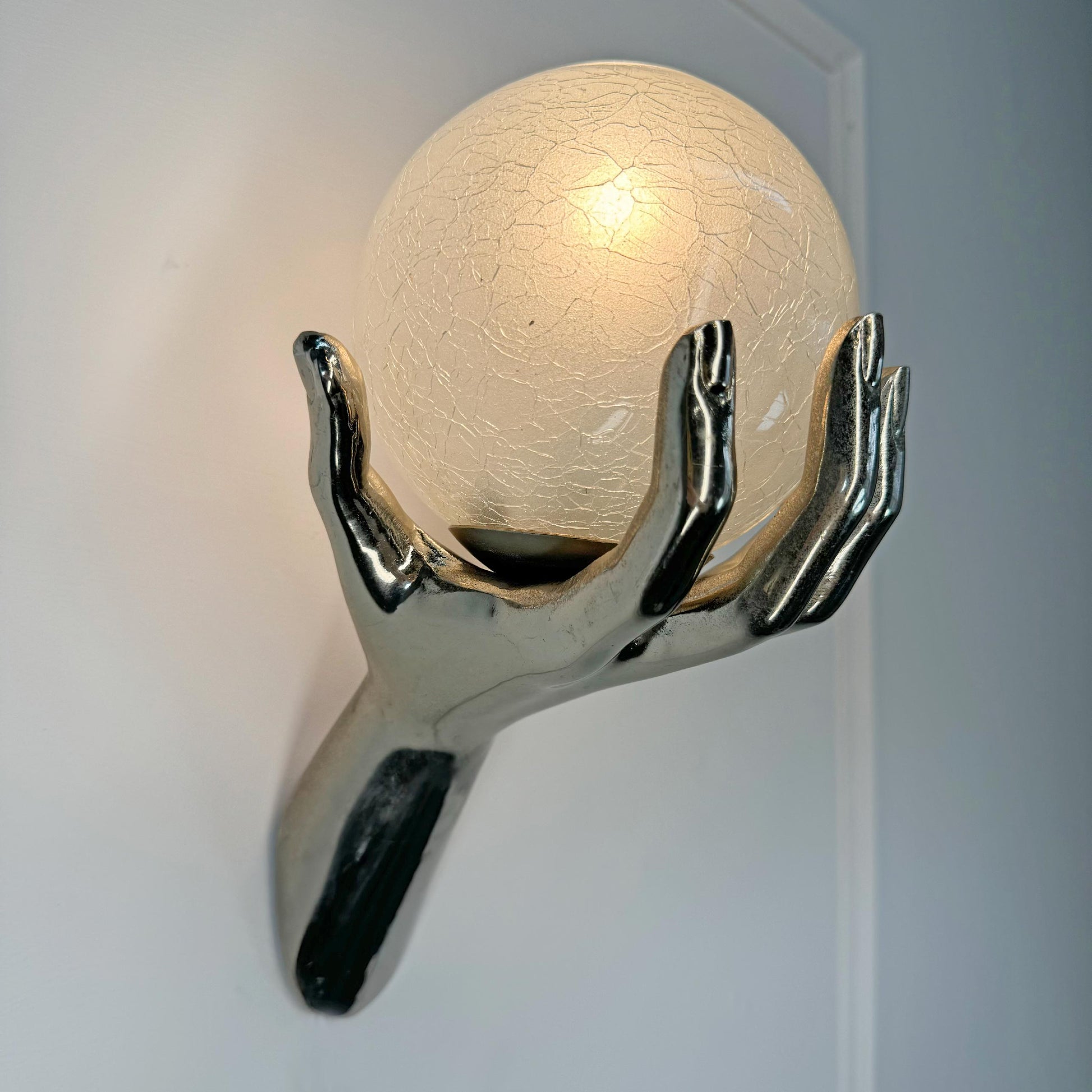 A sculptural maison arlus wall sconce featuring a glossy metallic hand holding a glowing, crackled glass orb against a plain light gray wall.