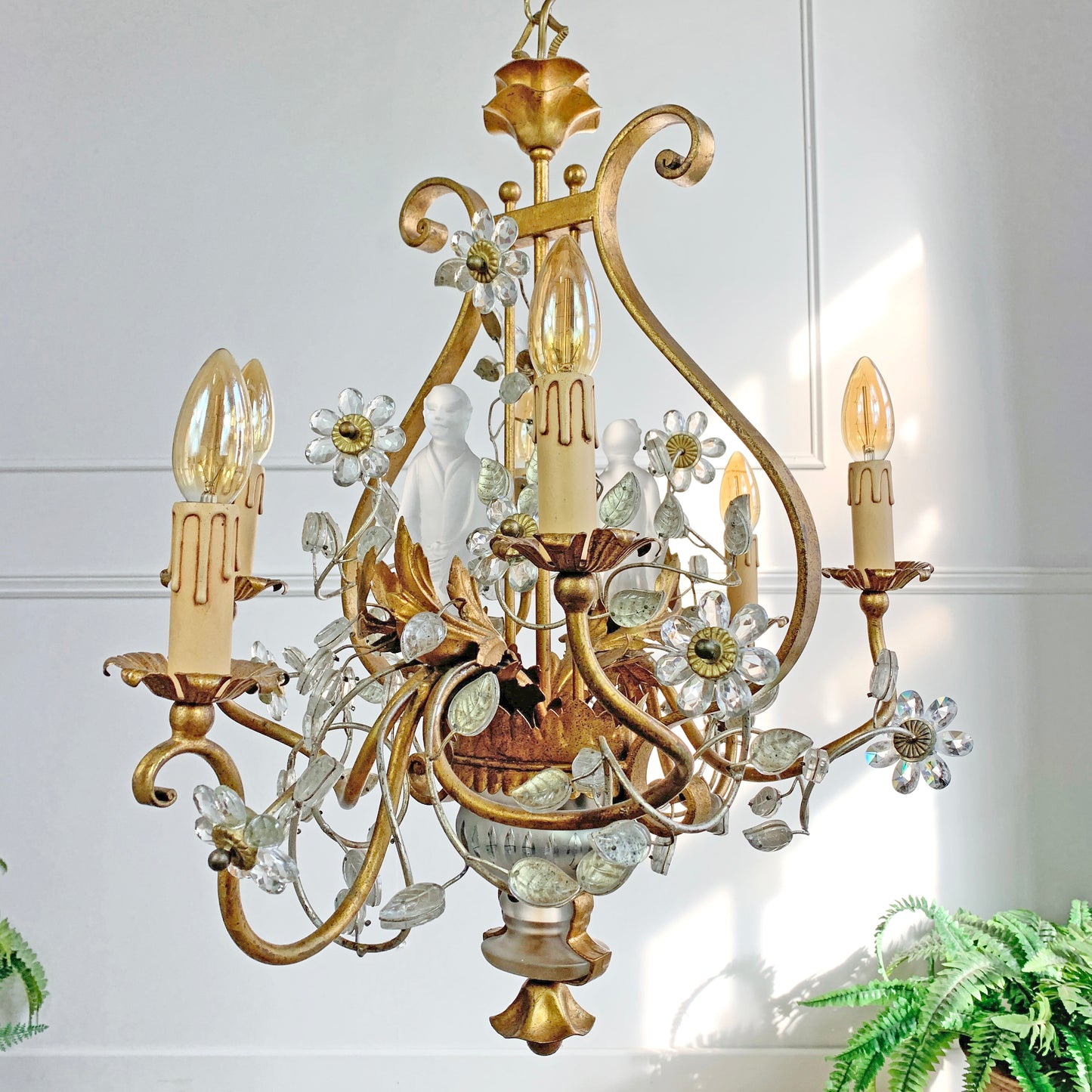 An ornate vintage Banci Firenze gold chandelier with candle-style lights and clear glass chinoiserie figures, shown hanging against a white paneled wall with sunlight streaming in.