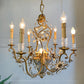 An ornate brass and crystal chandelier with six candle-style lights, clear crystal accents, and floral scrollwork, shown hanging against a light gray wall.