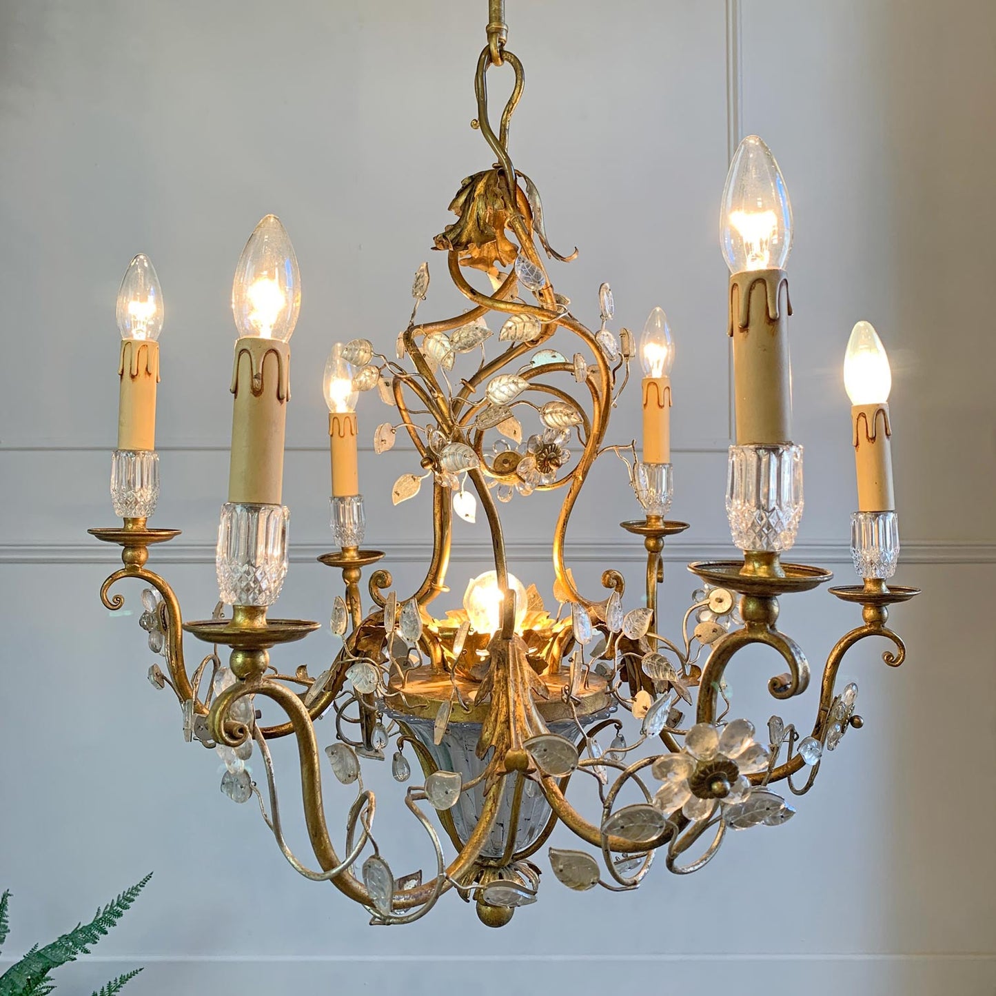 An ornate brass and crystal chandelier with six candle-style lights, clear crystal accents, and floral scrollwork, shown hanging against a light gray wall.