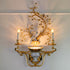 An ornate wall sconce with a gold-tone metal frame, featuring a central crystal bird figurine, floral branches with clear crystal flowers, and two lit candle-style bulbs on a white wall.