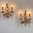 Two ornate designer wall sconces with gold-toned metal scrollwork, candle-style bulbs, and hanging clear crystal prisms, mounted on a plain light wall.