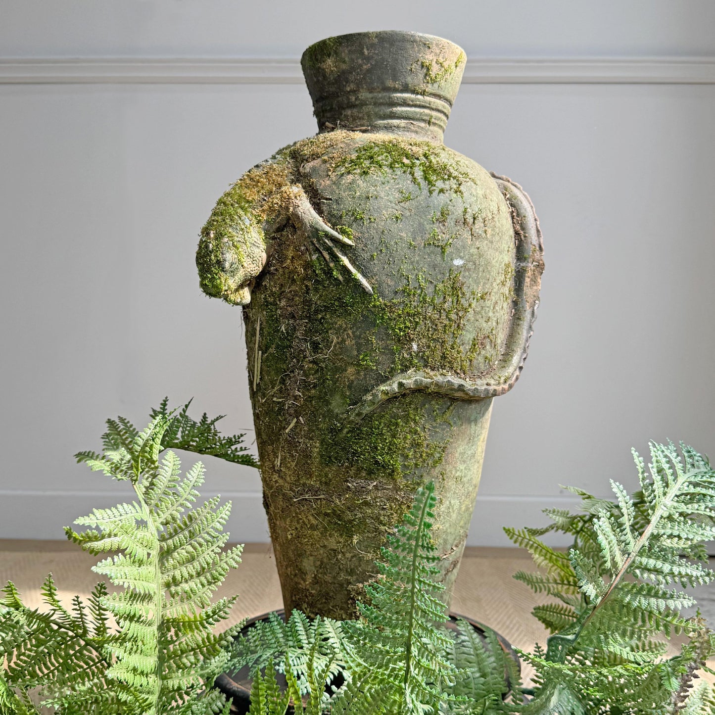 A mossy green terracotta urn with a lizard sculpted on ithe side, surrounded by green ferns in the foreground against a light gray wall.