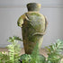 A mossy green terracotta urn with a lizard sculpted on ithe side, surrounded by green ferns in the foreground against a light gray wall.