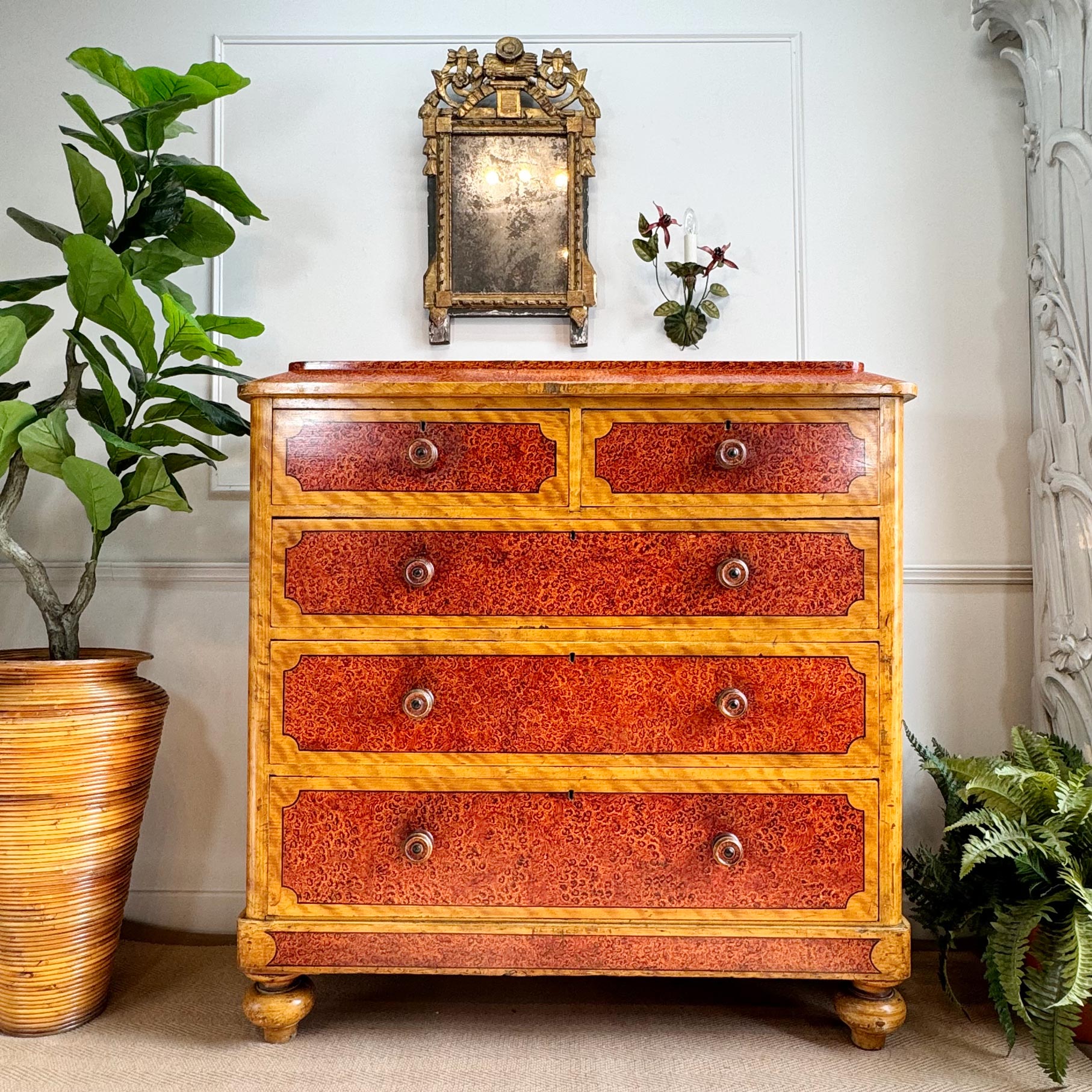 An ornate antique chest of drawers with red burl wood drawer fronts and a light wood frame, styled in a room setting with potted plants and a mirror.
