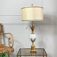 A table lamp with a cream fabric drum shade, a gold-tone metal pineapple-style stem and base, and a glossy white glass orb in the center, displayed on a glass-top side table against a white paneled wall.
