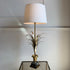 A table lamp with a large white drum shade, a brass-toned central stem, and sculptural metallic leaf-like branches rising from a decorative base, shown on a glass-topped table against a light neutral wall.