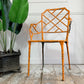An orange-painted metal garden chair with a lattice backrest and faux-bamboo legs, photographed indoors against a white paneled wall with a potted plant on the left.