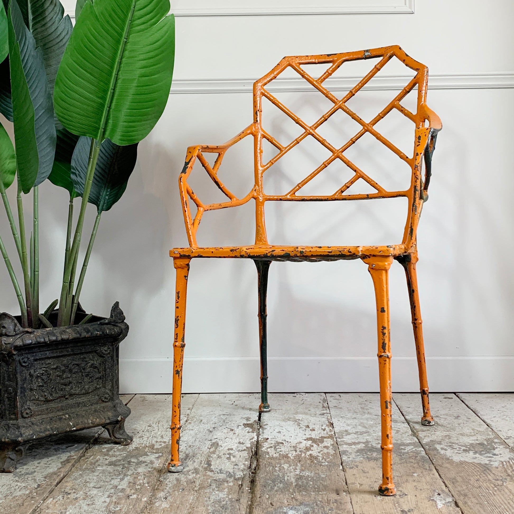 An orange-painted metal garden chair with a lattice backrest and faux-bamboo legs, photographed indoors against a white paneled wall with a potted plant on the left.