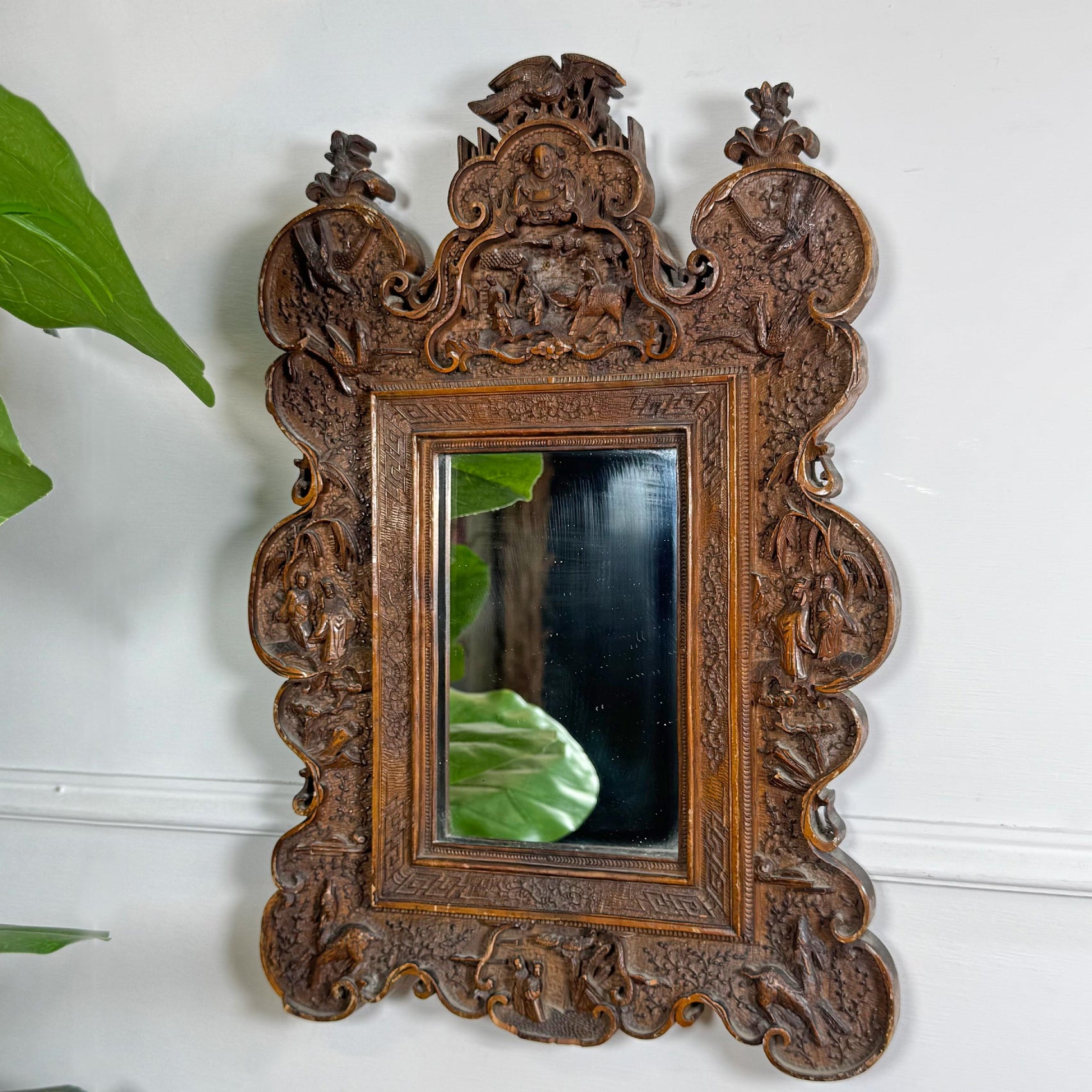 An ornate, asian carved wooden wall mirror with a highly detailed baroque-style frame, mounted on a white wall with a green plant leaf visible on the left.