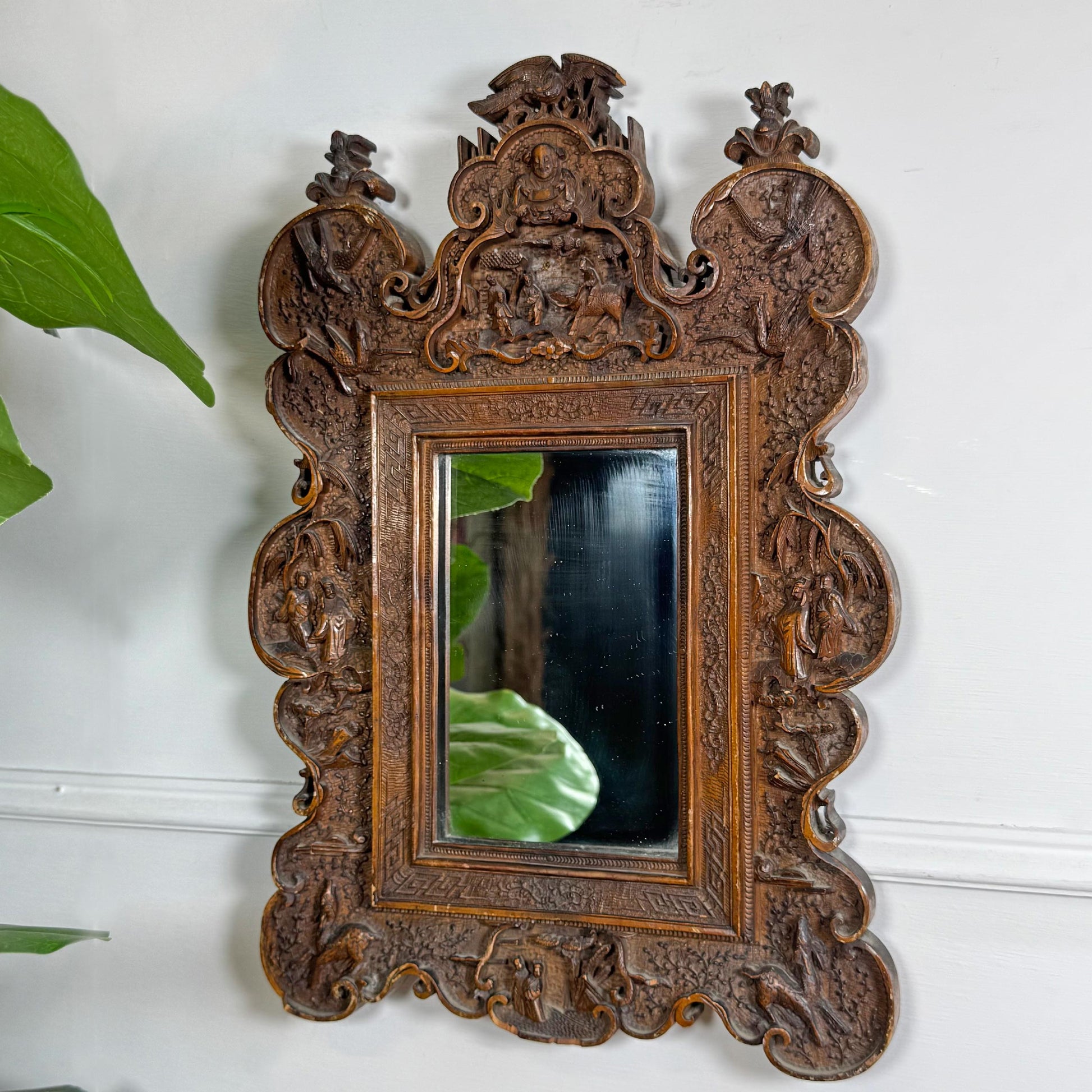 An ornate, asian carved wooden wall mirror with a highly detailed baroque-style frame, mounted on a white wall with a green plant leaf visible on the left.