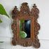 An ornate, asian carved wooden wall mirror with a highly detailed baroque-style frame, mounted on a white wall with a green plant leaf visible on the left.