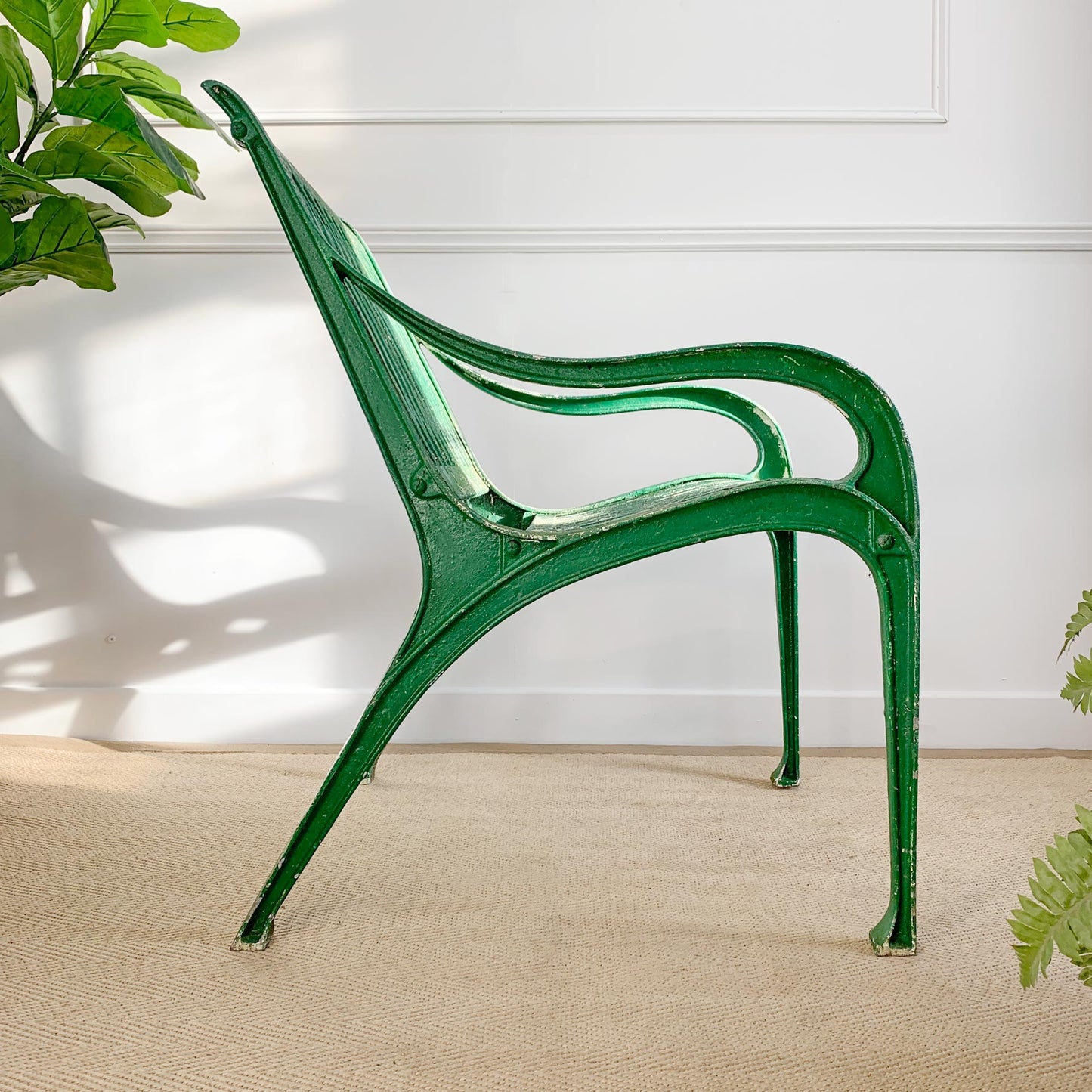 A green metal armchair with a distressed finish, positioned on a beige textured rug against a white paneled wall with soft shadows and green plants in the corners.