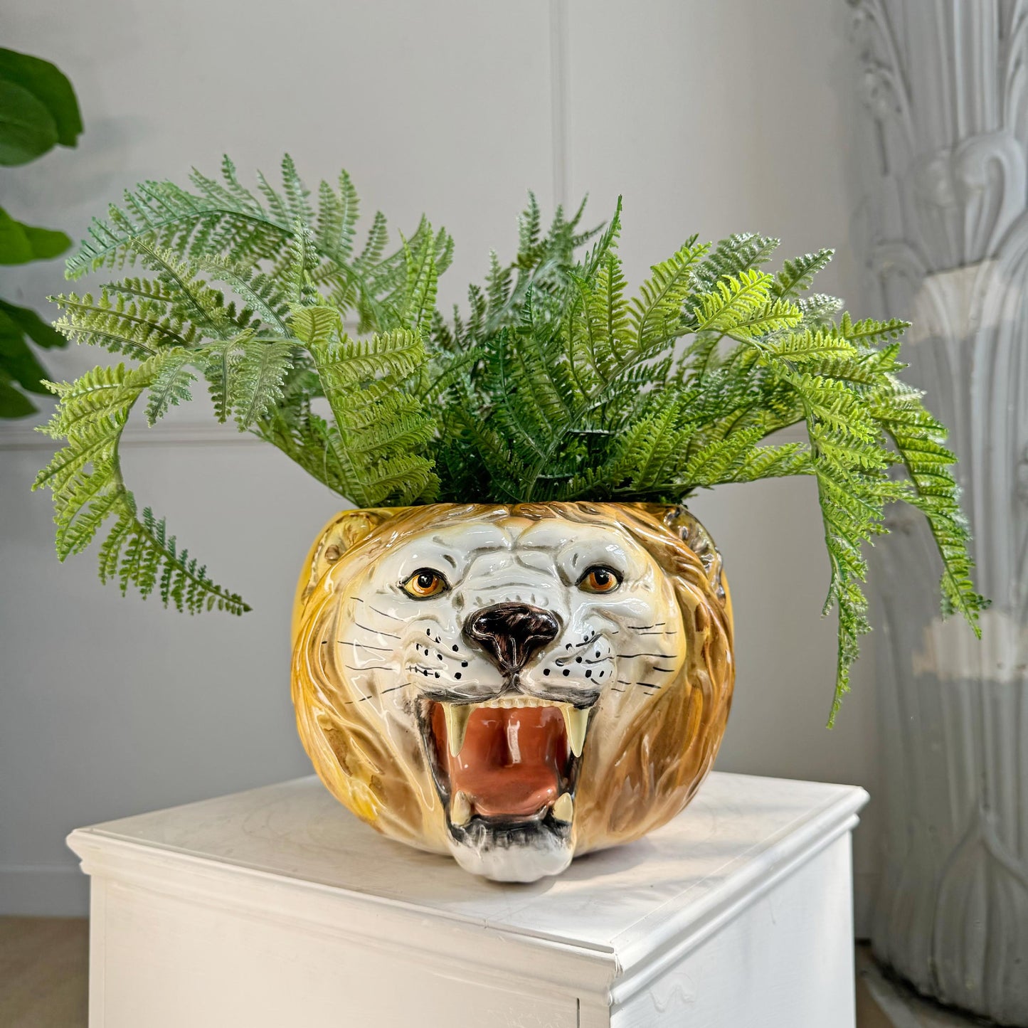 A vintage Italian Lion ceramic planter shaped like a roaring lion's face, filled with lush green ferns, displayed on a white pedestal in an indoor setting.