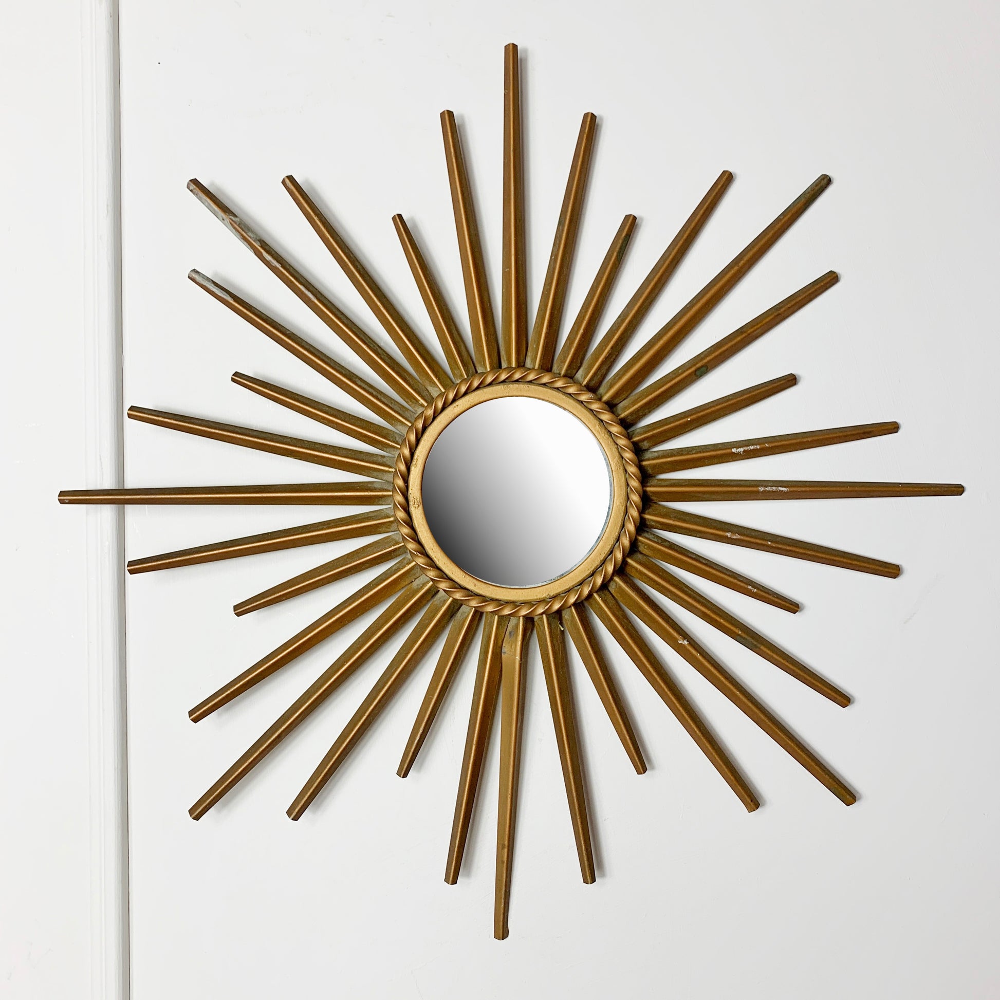 A Chaty Vallauris gold sunburst wall mirror with a circular mirror center and radiating metal rays, mounted on a plain white wall.