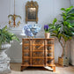 An ornate carved wooden chest of drawers in a warm medium brown tone, styled in a classic interior setting with a blue and white porcelain vase, a potted purple hyacinth, and lush green plants.