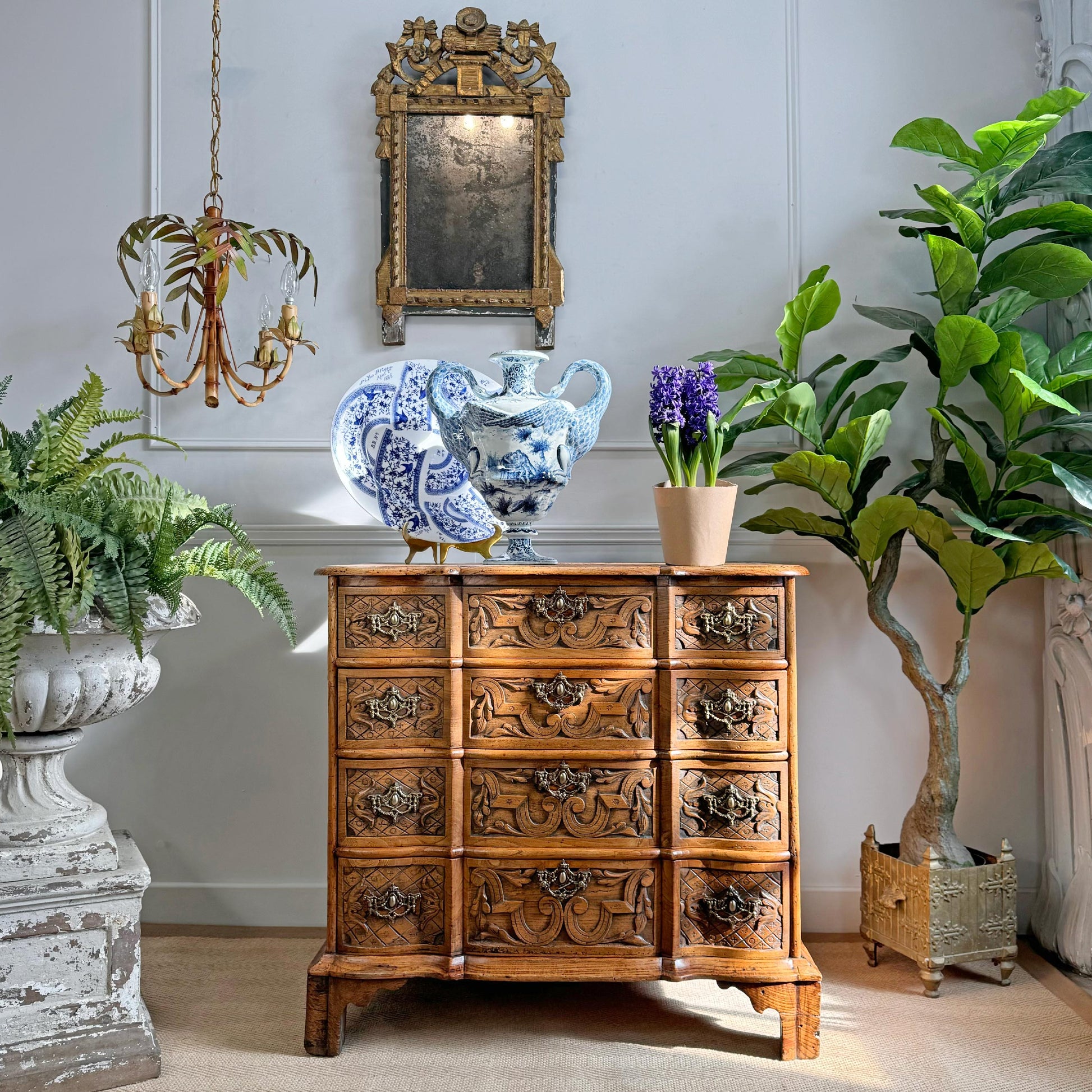An ornate carved wooden chest of drawers in a warm medium brown tone, styled in a classic interior setting with a blue and white porcelain vase, a potted purple hyacinth, and lush green plants.