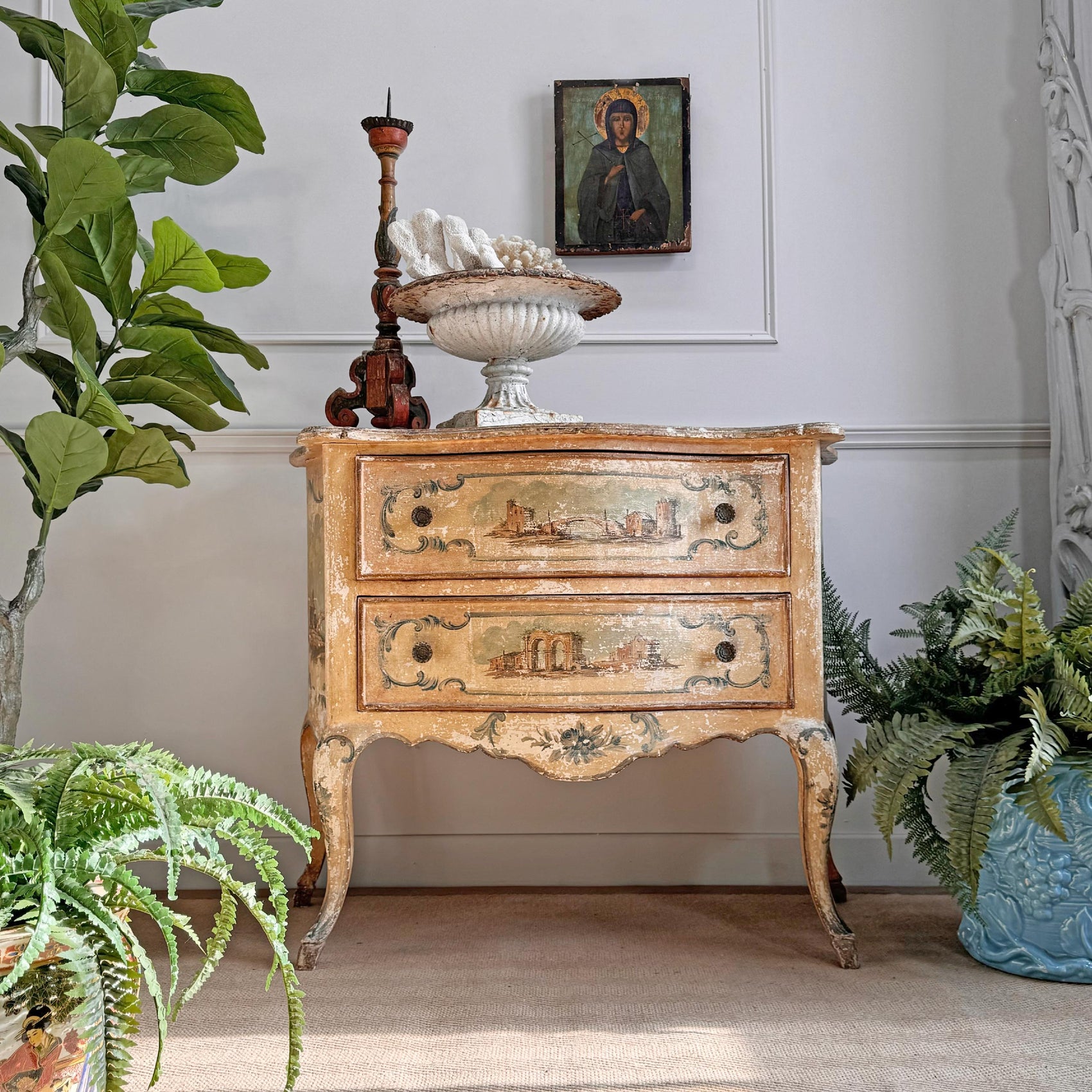 An antique venetian carved wooden chest of drawers with two drawers, featuring a distressed beige and gold finish with hand-painted landscape motifs. The chest is styled in a room setting with potted plants, a white seashell bowl, and a small religious icon on top.