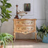 An antique venetian carved wooden chest of drawers with two drawers, featuring a distressed beige and gold finish with hand-painted landscape motifs. The chest is styled in a room setting with potted plants, a white seashell bowl, and a small religious icon on top.