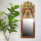 An ornate vintage chinoiserie wall mirror with a highly decorated, hand-painted frame featuring a beige background, red borders, and small painted figures, shown hanging on a white panelled wall next to a green potted plant.