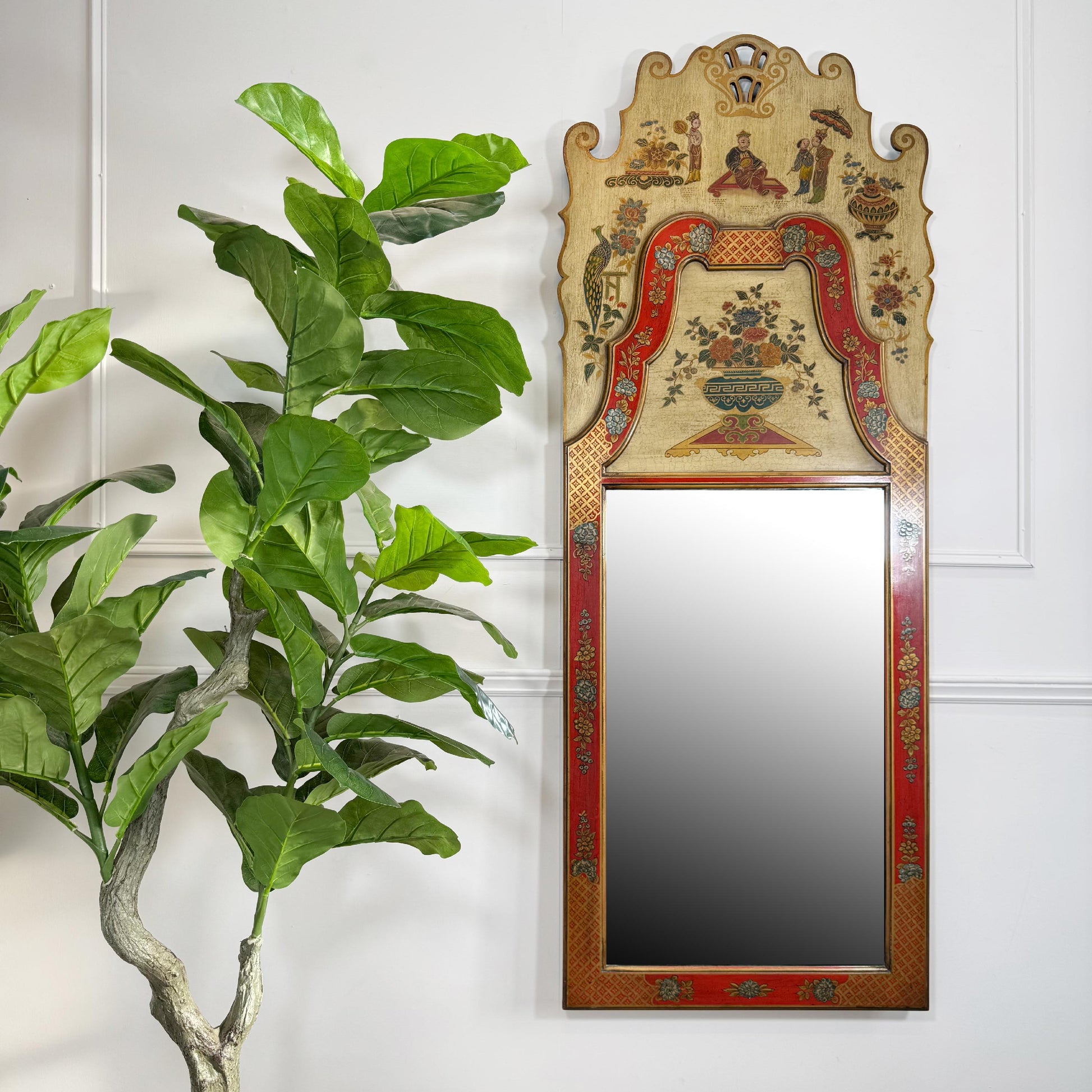 An ornate vintage chinoiserie wall mirror with a highly decorated, hand-painted frame featuring a beige background, red borders, and small painted figures, shown hanging on a white panelled wall next to a green potted plant.