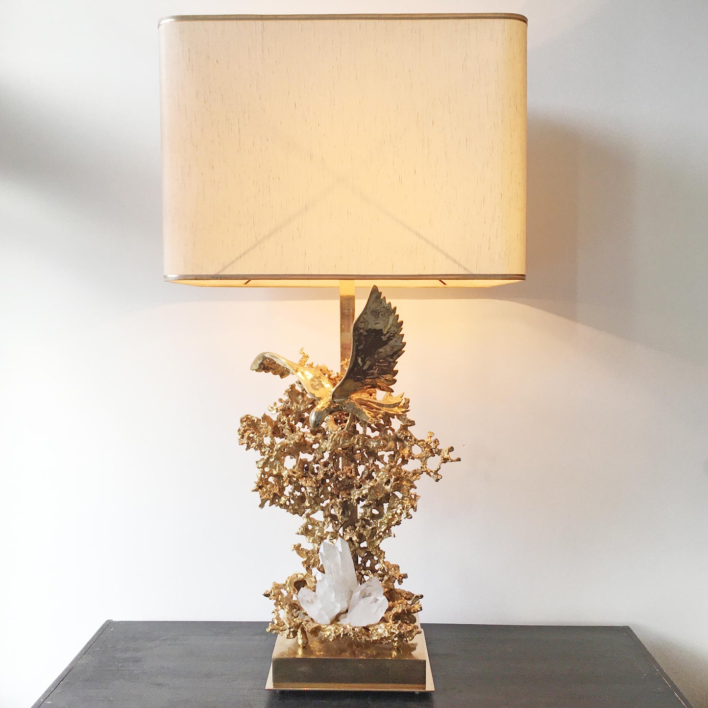 A decorative table lamp with a large beige fabric drum shade, a gold-plated sculptural base featuring an eagle and coral-like branches, and a white crystal accent, shown on a dark tabletop against a light wall.