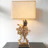 A decorative table lamp with a large beige fabric drum shade, a gold-plated sculptural base featuring an eagle and coral-like branches, and a white crystal accent, shown on a dark tabletop against a light wall.