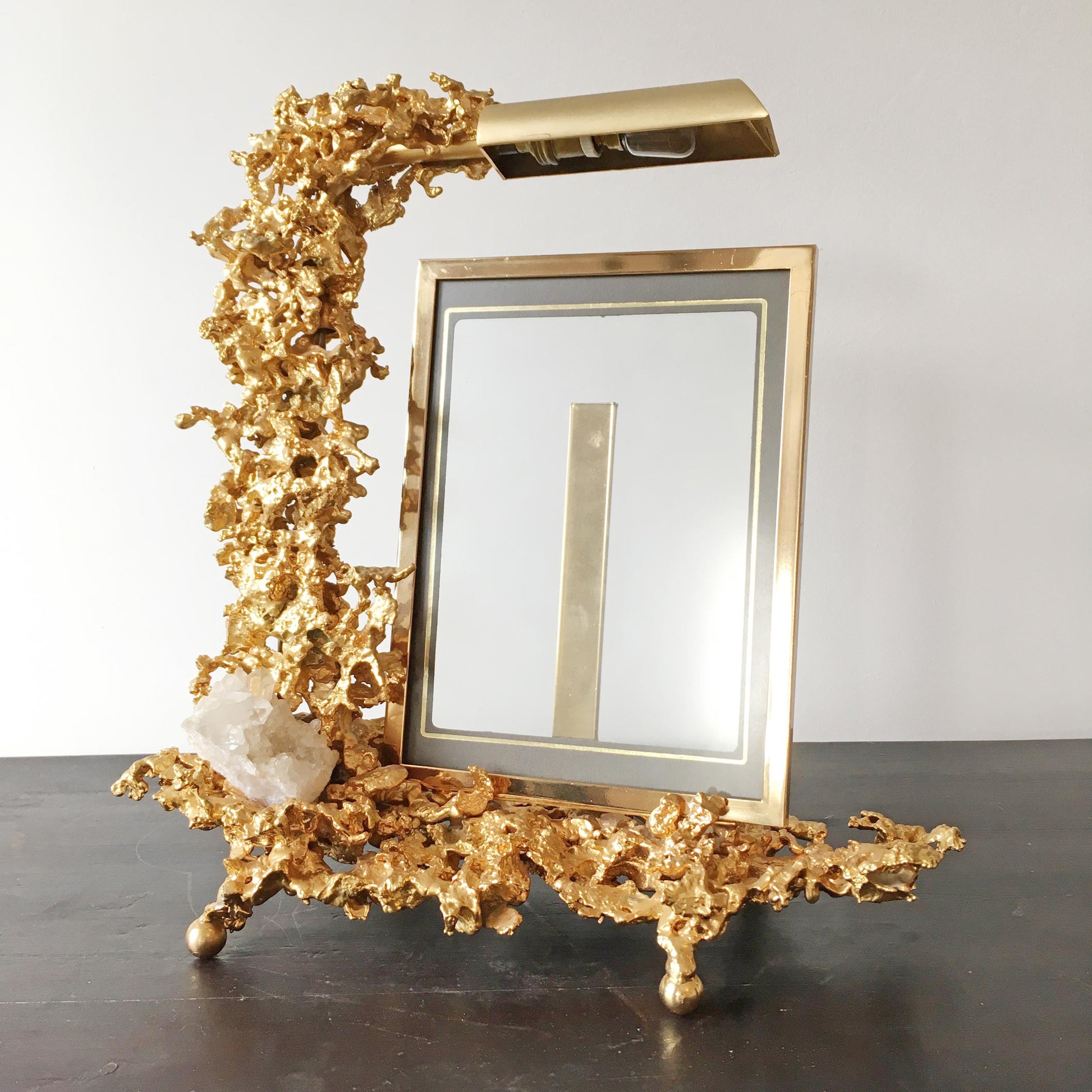 A decorative gold-plated designer picture frame stand with an ornate coral-like base, a matching top arm, and a small clear crystal accent, photographed against a plain light background.