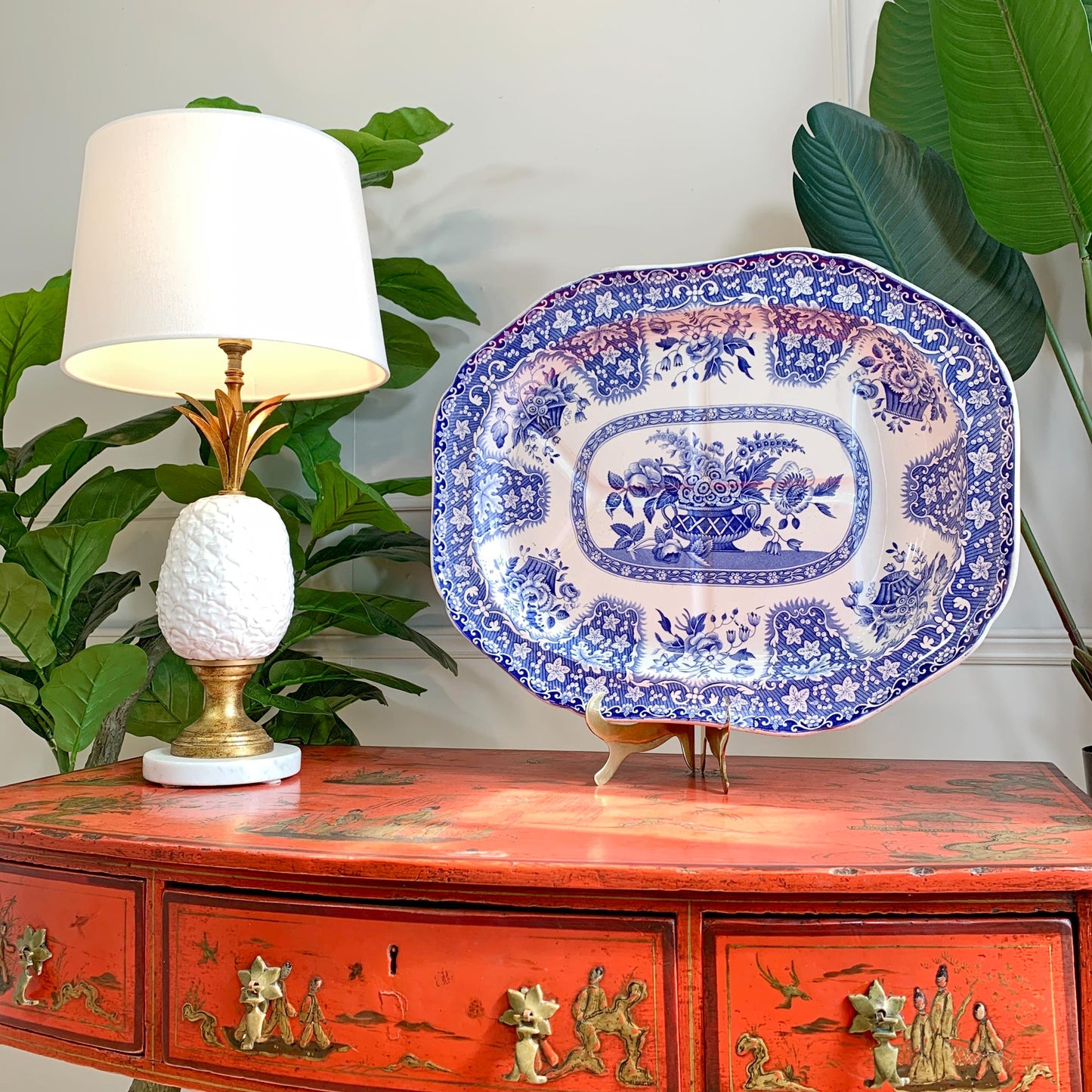 A blue and white porcelain serving platter with floral patterns, displayed on a small stand on top of a distressed red wooden console table next to a lit table lamp with green plants in the background.