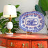 A blue and white porcelain serving platter with floral patterns, displayed on a small stand on top of a distressed red wooden console table next to a lit table lamp with green plants in the background.