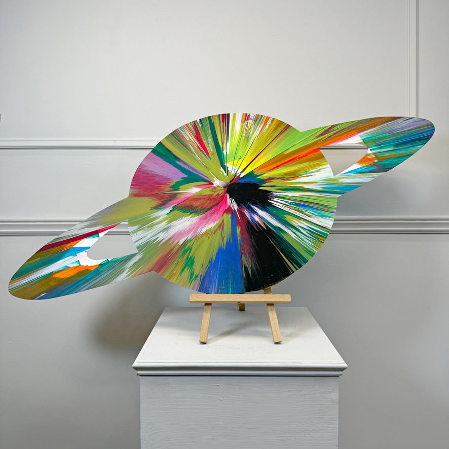 A Damien Hirst colorful abstract ringed planet sculpture made of painted wood, displayed on a small wooden easel stand atop a white pedestal against a light gray paneled wall.